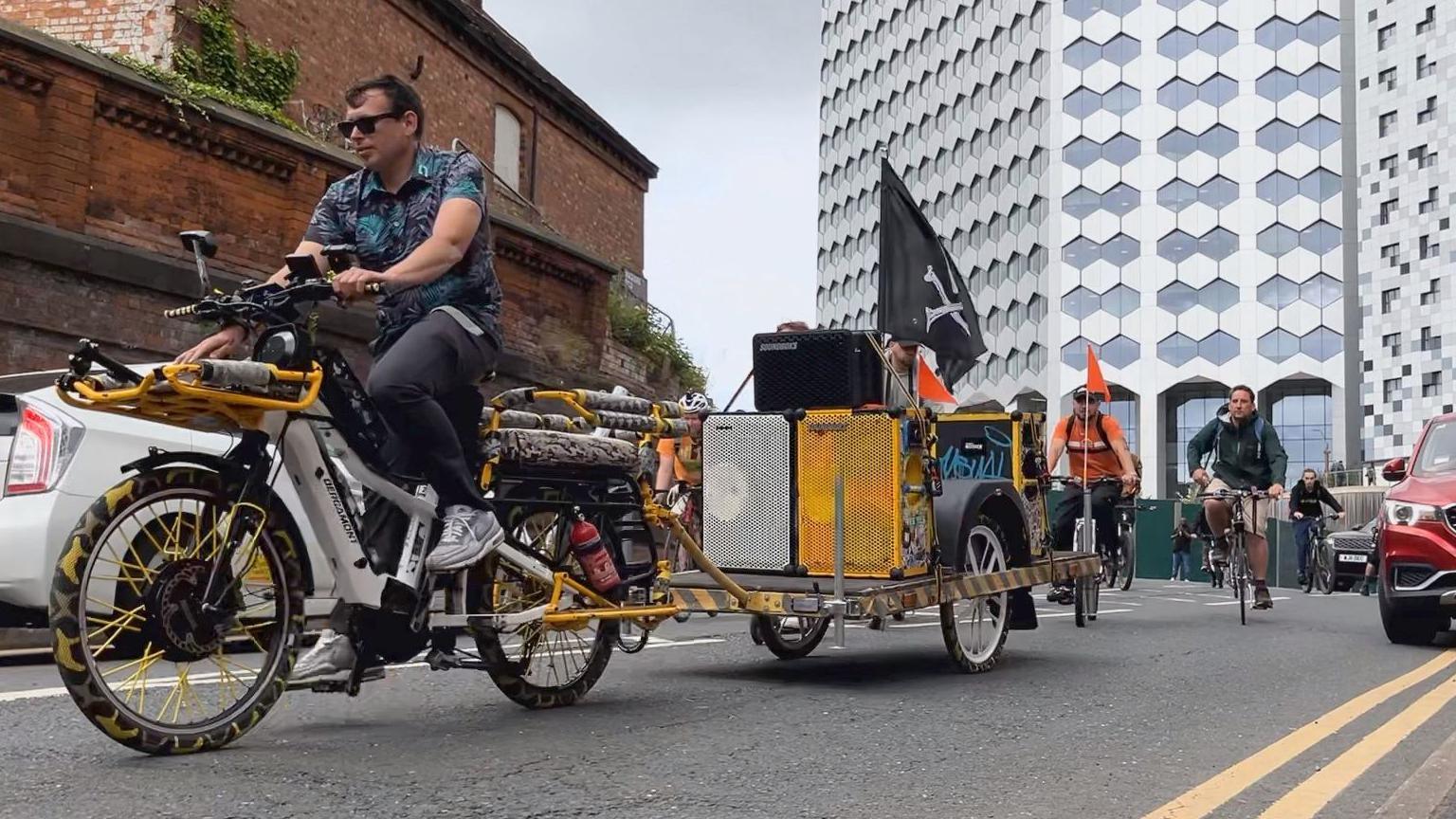 A man is cycling on a white bike with a trailer carrying large music speakers. There is a black flag on the back of the bike with a skull.