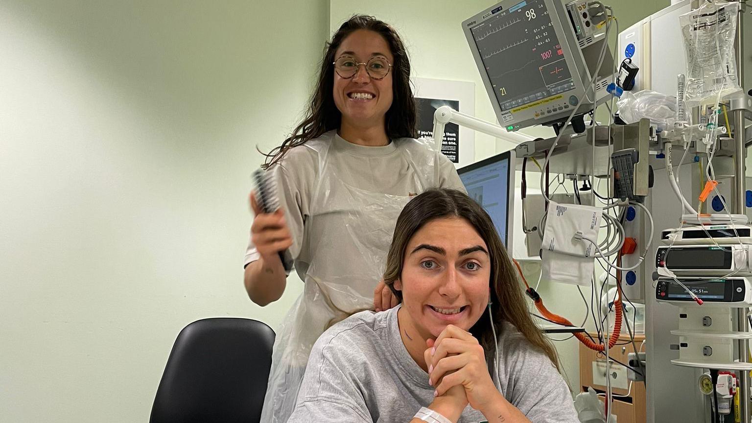 Emma Wassell is smiling at the camera while sitting on her hospital bed, while a friend brushes Emma's hair behind her