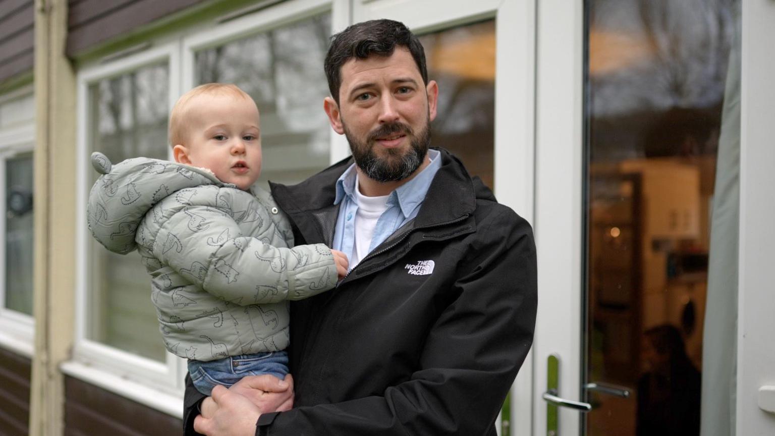 Tom standing outside his flat on a cold winter's day holding his little boy in his arms