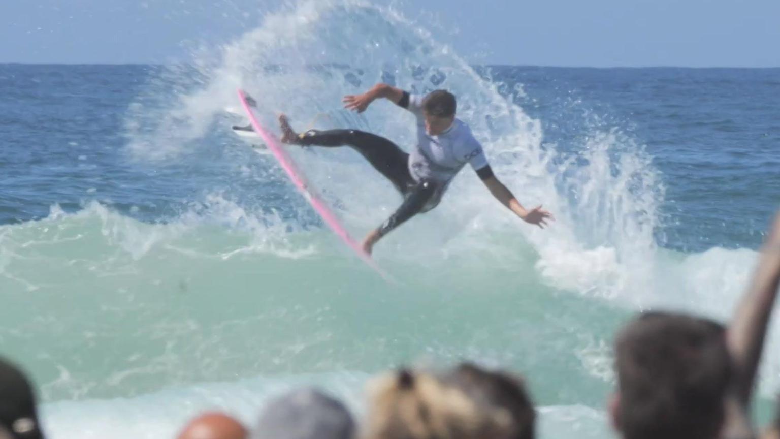 Lukas Skinner is in the air on his pink surfboard with the sea and wave spray all around him - he is wearing a Boardmasters white rash vest and a black wetsuit and you can see the heads of spectators in the foreground