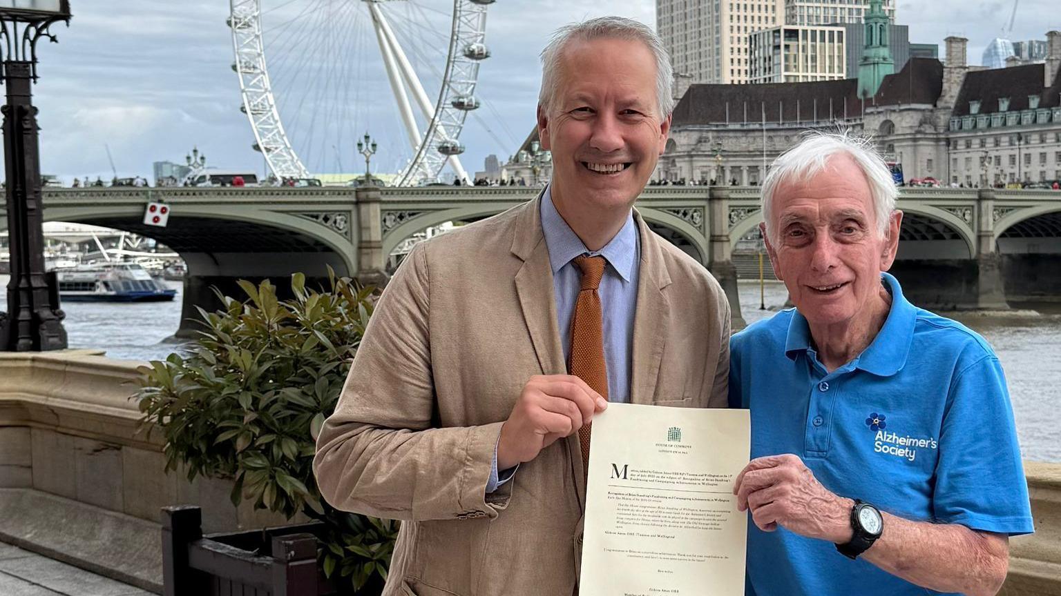 An older man in a blue Alzheimer's Society T-shirt is standing next to a man in a brown suit jacket. In the background is the London Eye.