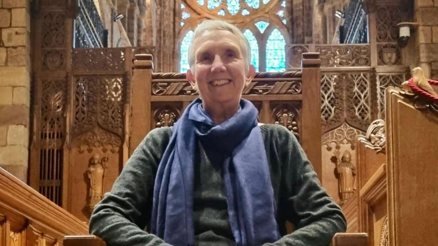 Ann Cleeves, sits on a wooden chair inside what looks like a church. She is smiling and wearing a green jumper and purple scarf.