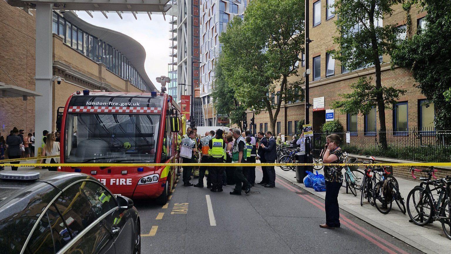 Emergency services are gathered by a fire vehicle outside a modern hospital building. There is tape closing off the road. 