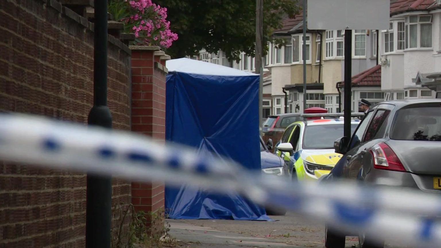 A blue tent is set up on Monks Park road. In front of it is a police cordon. Next to the tent is a parked police car. Houses can be seen in the background. 