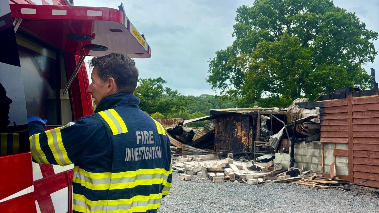 A male firefighter stands in front of a section of a heavily damaged building. He is wearing a blue, yellow and silver jacket, which says "FIRE INVESTIGATION" on it in silver capital letters. Behind him, there is a pile of bricks on the ground in front of a collapsed wall.