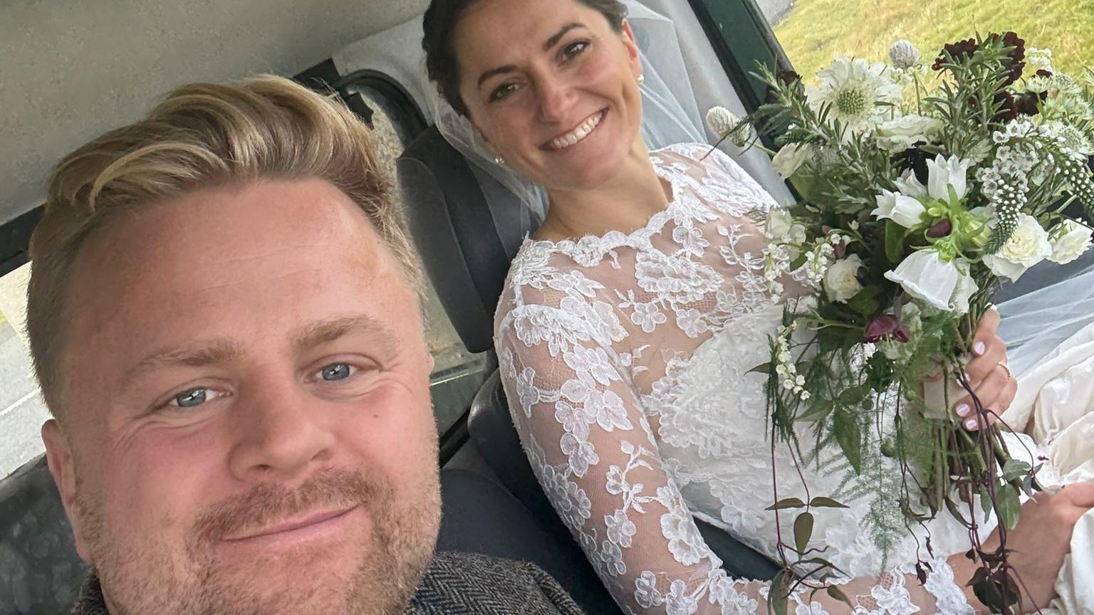 A man and a woman sit inside a vehicle, dressed for a wedding. On the left, a man in a brown tweed suit with a maroon tie smiles warmly. On the right, a woman in a white lace wedding dress holds a bouquet of white and green flowers, also smiling. The background shows a rural road and grassy landscape through the vehicle windows.