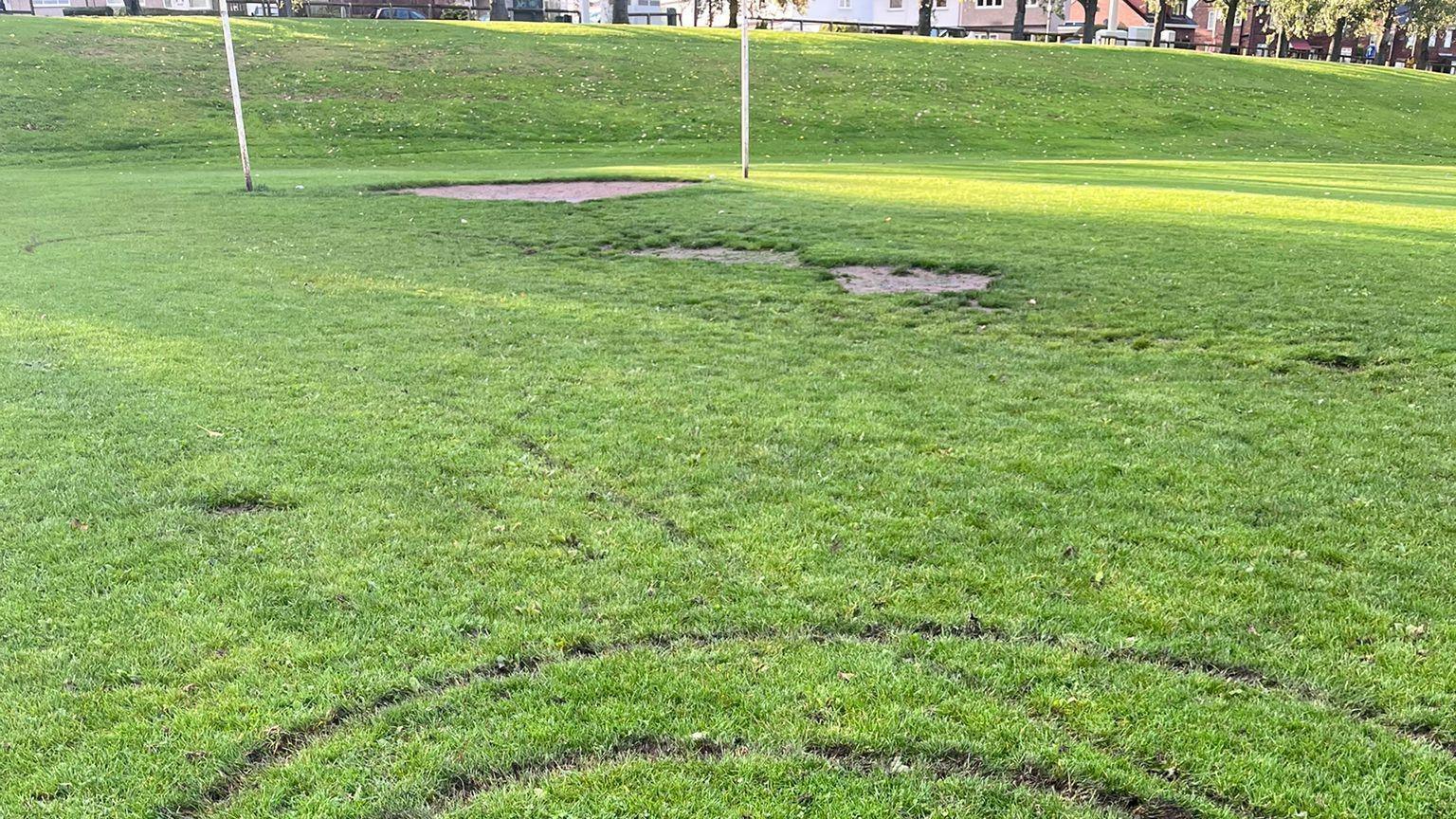 The image shows a football pitch and in the foreground you can see tyre tracks, behind is a goal mouth. 