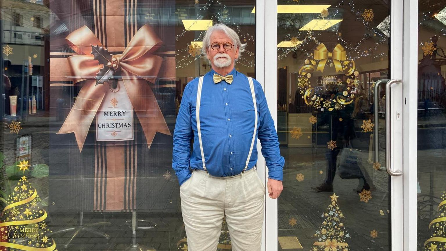 Charles McEleney stands outside the hairdressers that he owns in Derry. He is wearing a blue shirt, and white trousers. he is also wearing a pair of white braces and a yellow bow tie