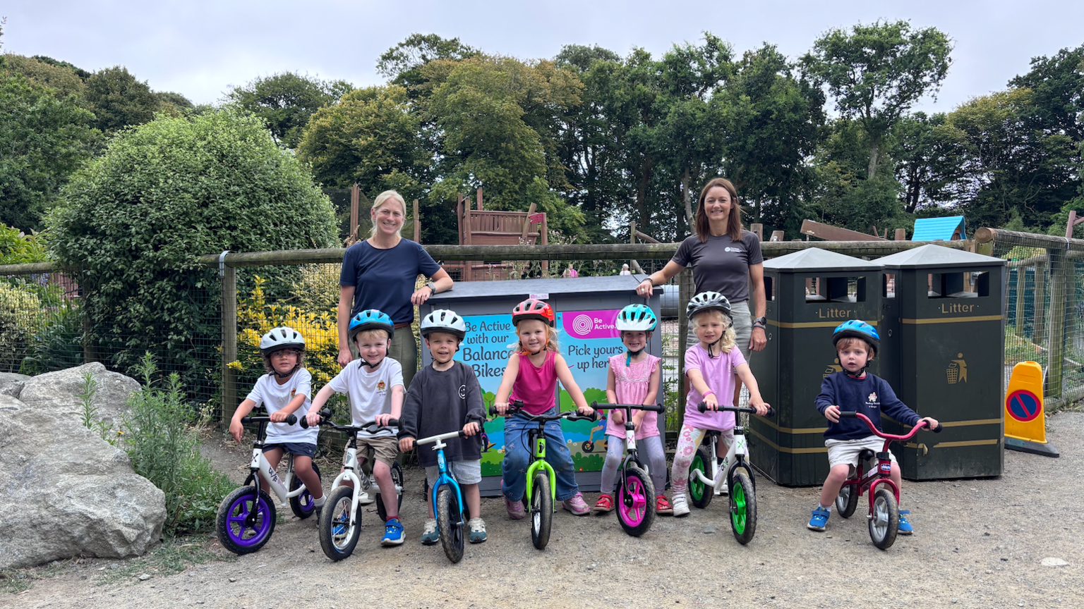 A row of children about age four and five on bikes, two women standing behind, at a playground. Trees and blue skies seen behind.