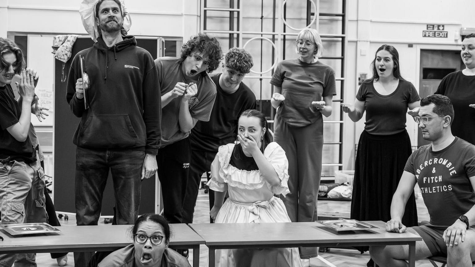 10 actors rehearsing a scene from The Little Mermaid. One is on the floor pulling a face, a woman looks shocked, with her hand on her mouth, one man is looking out with a hat, the rest are looking down at the women in shock. The image is black and white and they are in a school hall with equipment behind on the wall. 