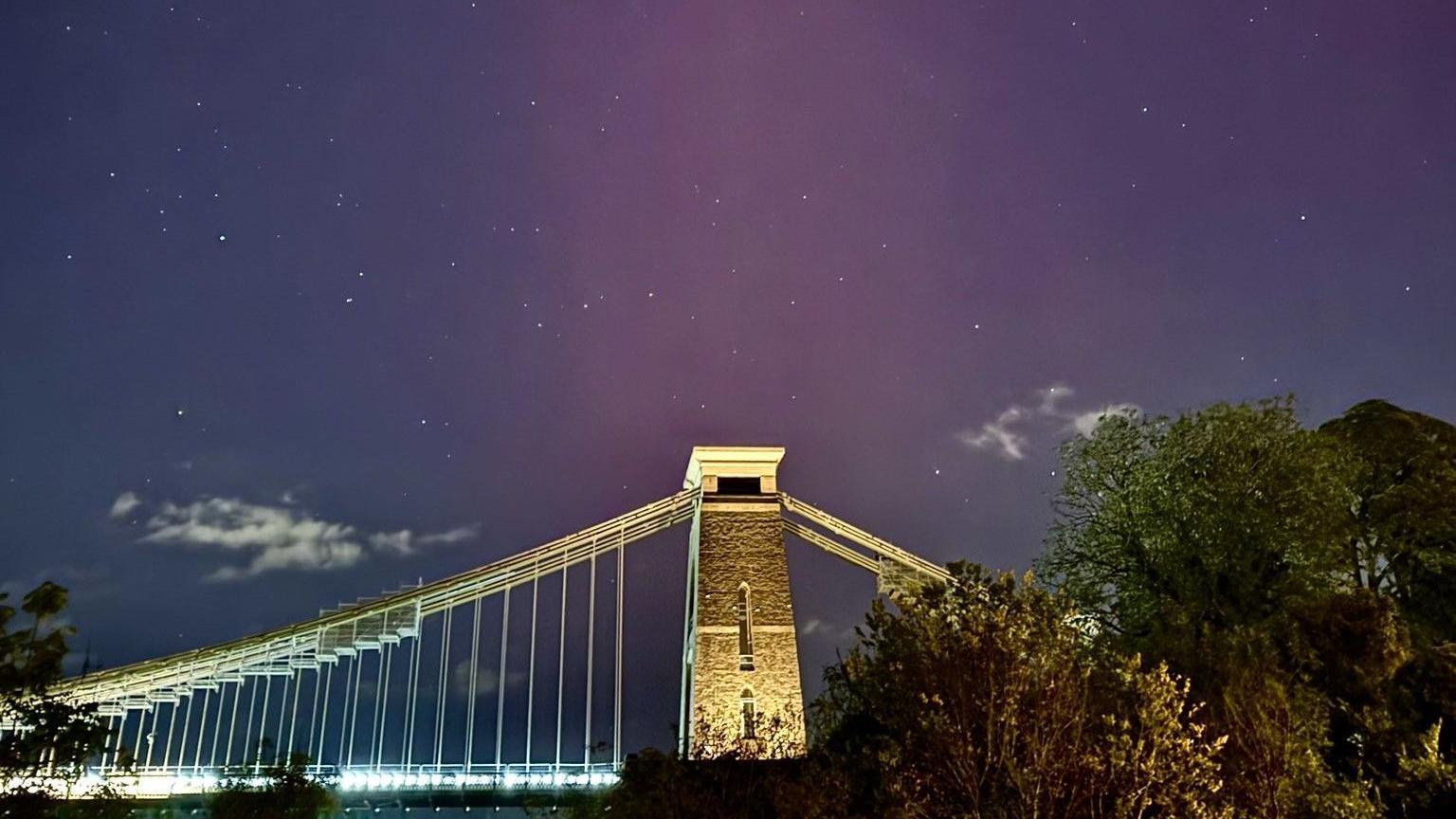 The Northern Lights are visible over the Clifton Suspension Bridge, which is photographed from below at night. The sky is a light purple and green