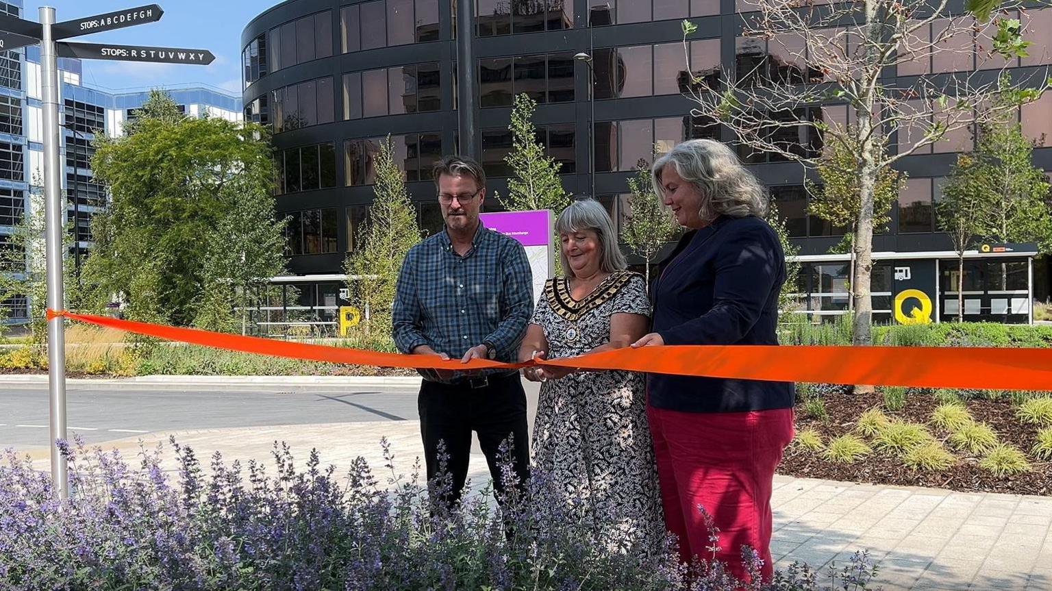 Three people stand behind a large red ribbon.  Councillor Chris Watts, mayor Fay Howard and MP Heidi Alexander are getting ready to cut the ribbon. They are standing in a public area, with freshly planted trees and flowers, clean pavements and fresh tarmac. A large building with glass windows is behind