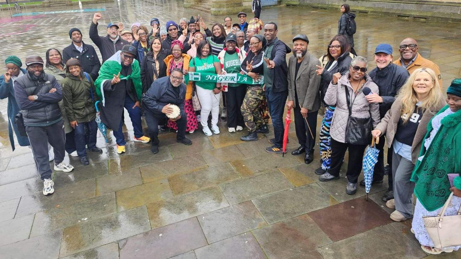 A line-up of people including some wearing green and white T-shirts and one man draped in a green and white Nigerian national flag. 