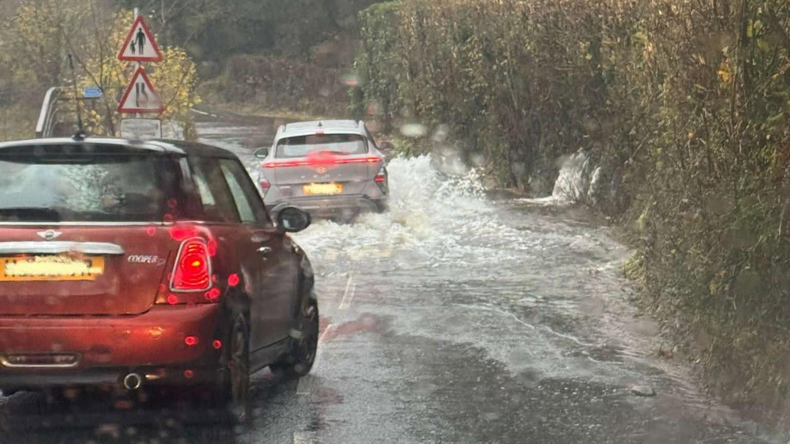 A flooded road at Newby Bridge, Cumbria. There two cars - one red and one grey - that are travelling down the road where there is a significant amount of water covering the road.
