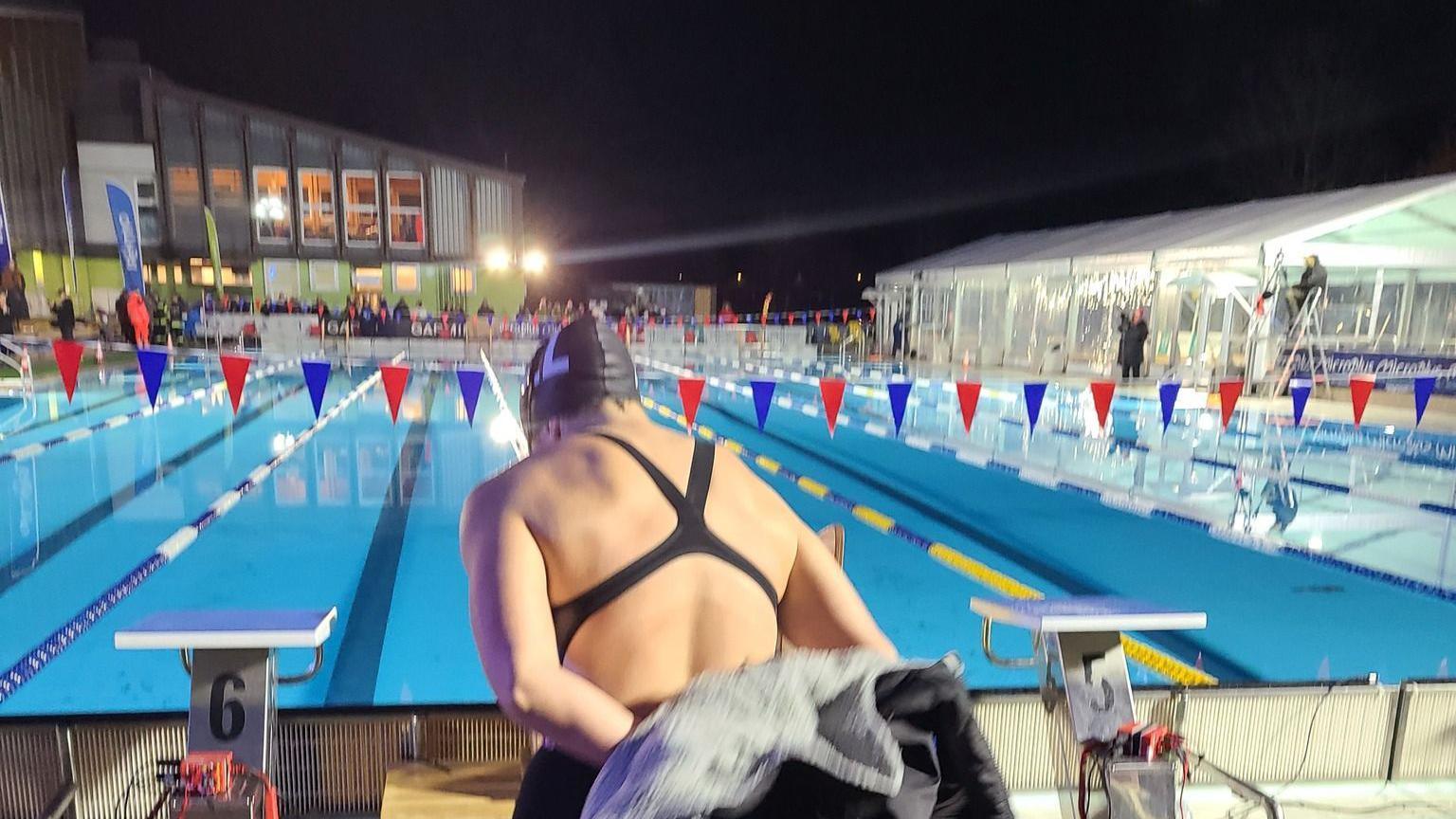 A swimmer disrobing ready to get into the pool. It is dark and there is some floodlighting.