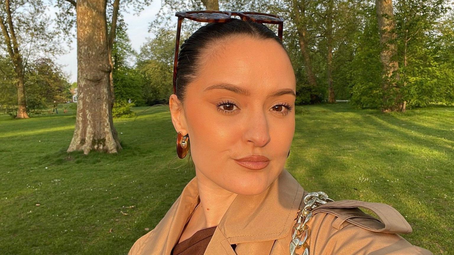 A selfie of a young woman in a park. She has brown hair pulled back and tortoiseshell sunglasses on her head. She has a light brown mac on  and dark brown hooped earrings. Trees and grass are in the background.