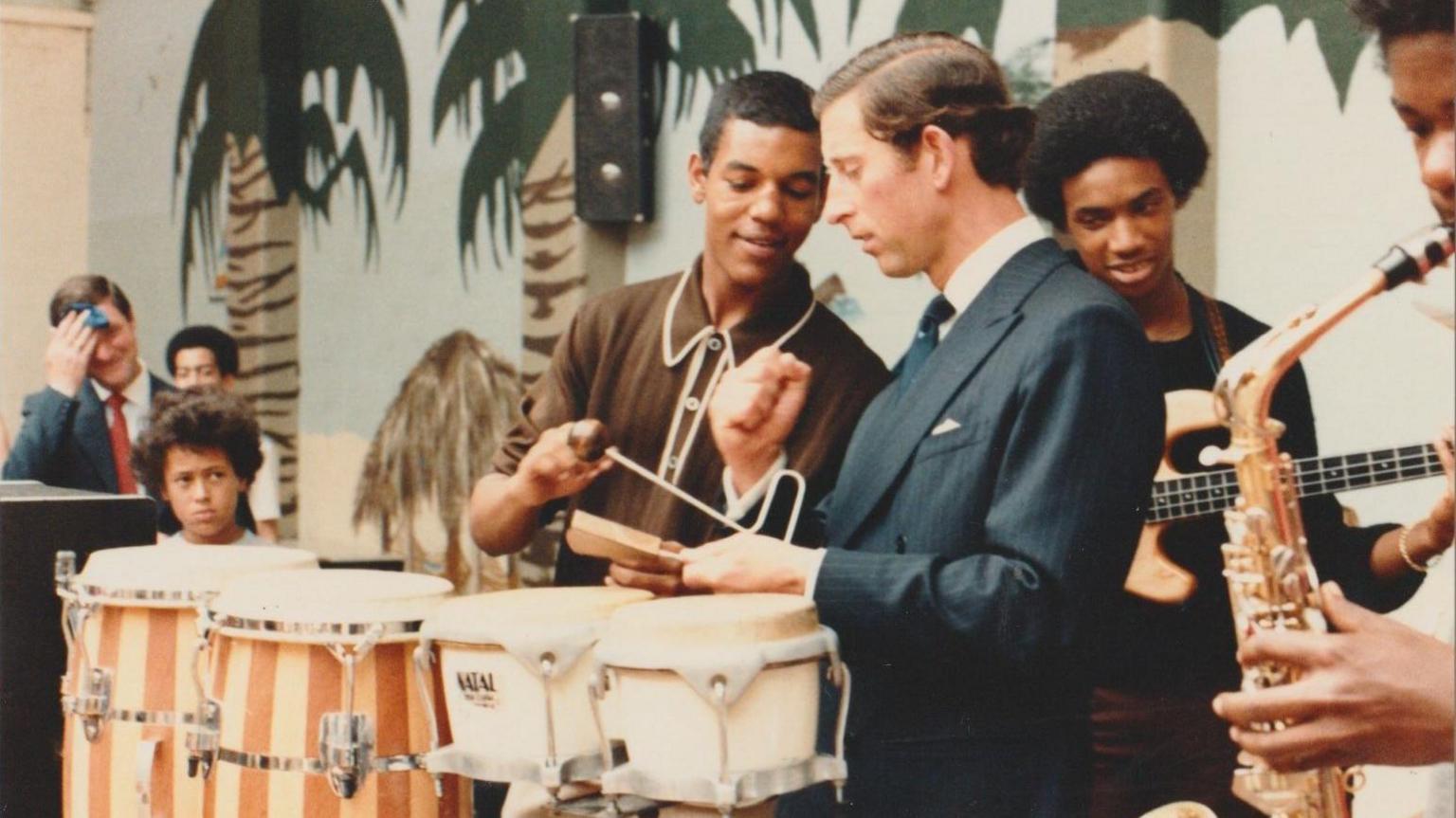 King Charles III stands behind some bongos with men playing saxophone and bass guitar in the background. A backdrop has palm trees painted on it. On the left, a boy is watching and a man in a jacket and tie is mopping his brow with a handkerchief.