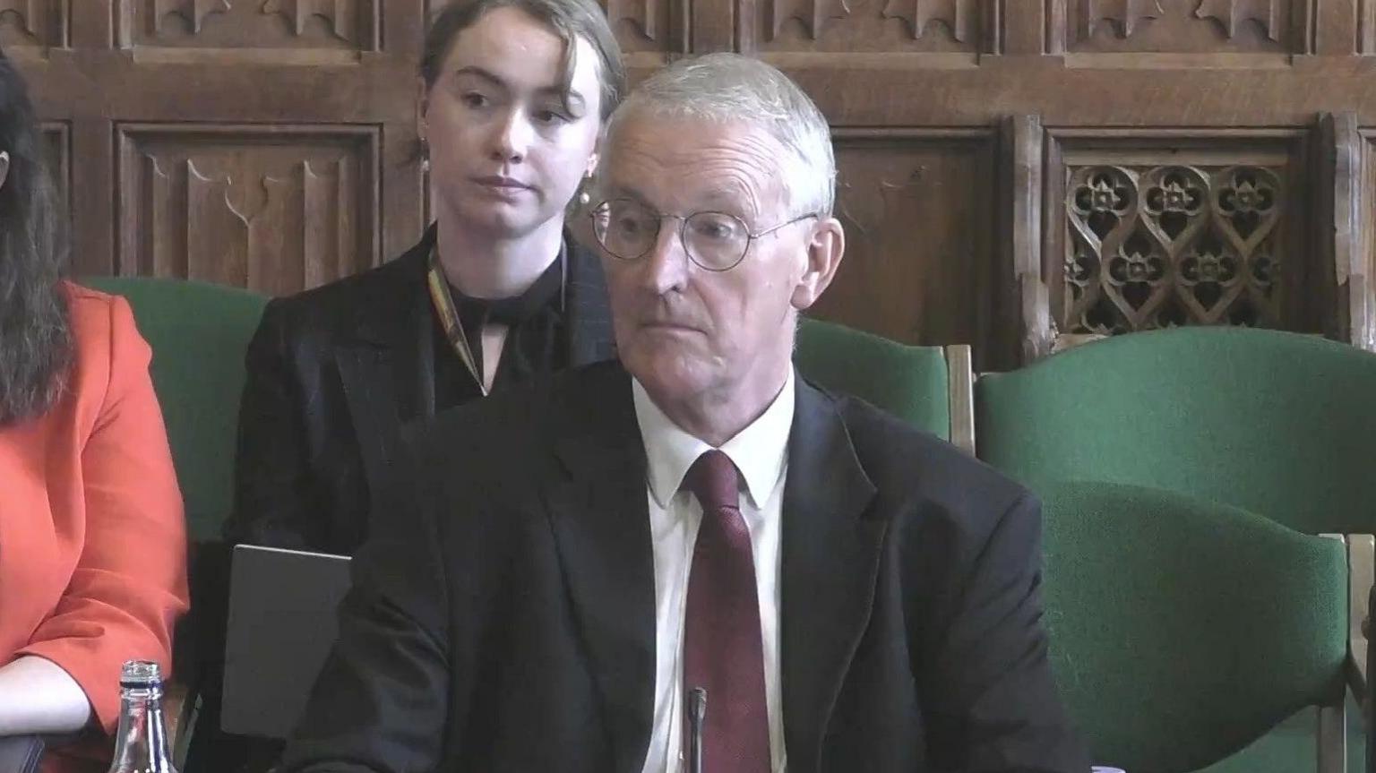 A still of Hilary Benn appearing at the Northern Ireland Affairs Committee in Westminster. He, as well as other attendees, are sitting in green fabric chairs. Benn is an older man with grey/white short hair and a pair of circular metal glasses. He is wearing a black suit jacket, a red tie and a white collared shirt