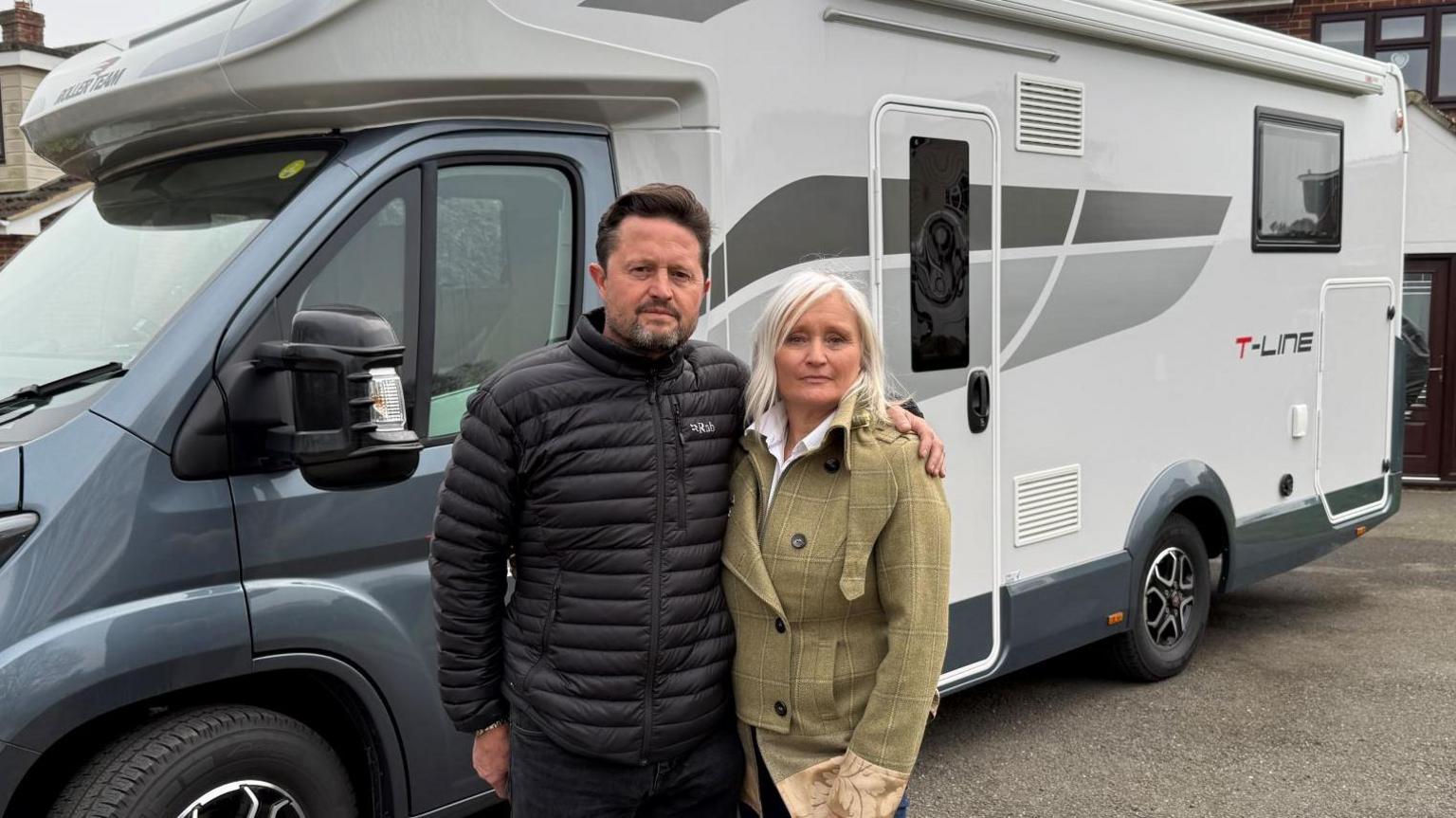 Adrian and Joanne Fenton wearing coats and standing in front of their navy and white motorhome, which is parked in front of their house. They have serious expressions on their faces.