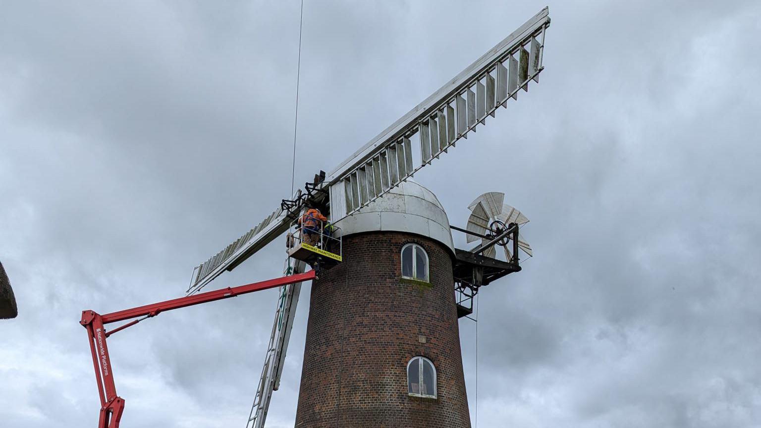 Under a dark grey sky, the top of the windmill, which is a round red brick building with a white dome on top, has a cherry picker lifting men to the white sails as they remove one