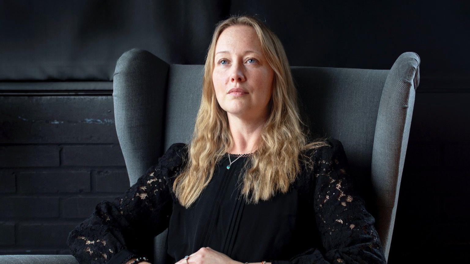 Katie Ward sits in a large grey armchair looking out of frame. She is blonde and wears a black top.