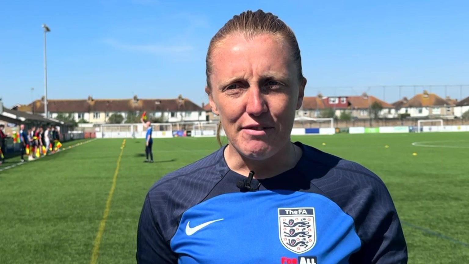 Head and shoulders image of Lisa Benn wearing a black and blue referee top in front of a training session on a football pitch
