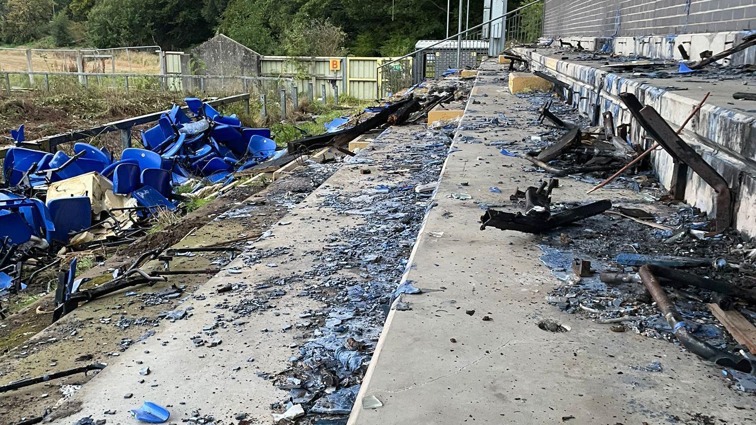 Shards of glass and debris litter a stand in a football ground.
A number of blue seats lay at the bottom of the pitch.