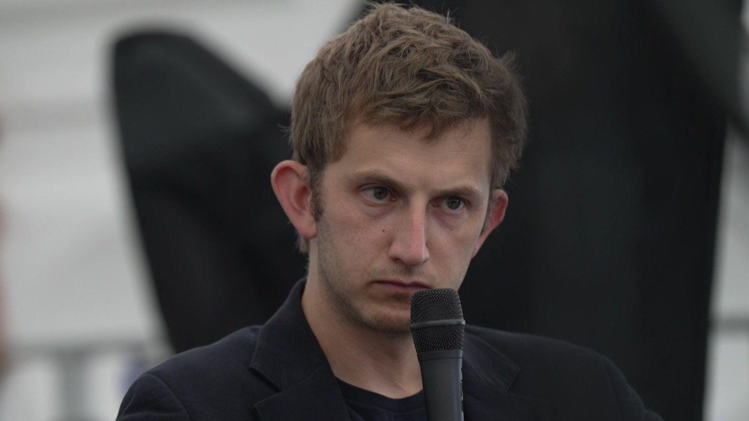 Owain Williams wearing a black jacket and holding a microphone to his face
