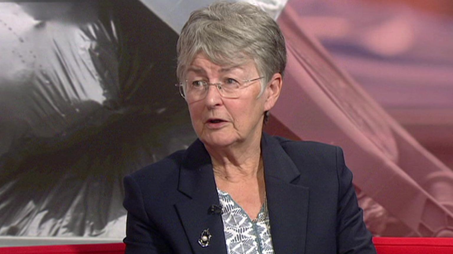 A woman with short grey hair, sitting on a red sofa in a television studio. She is wearing a navy blazer and blue and white top.