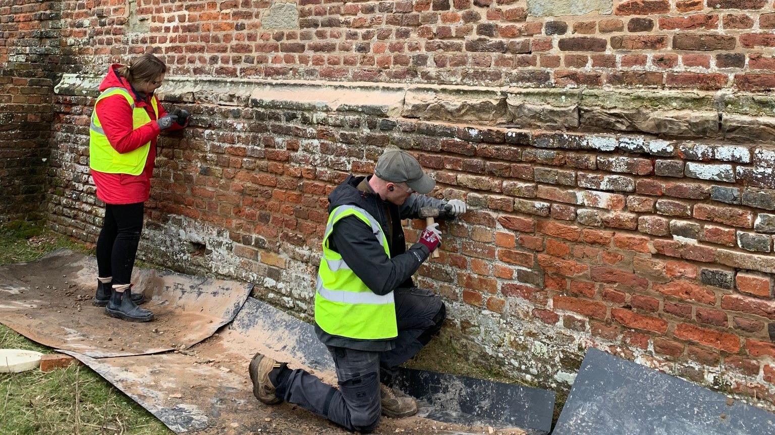 Two people wearing high-vis jackets working on a red brick wall with tools