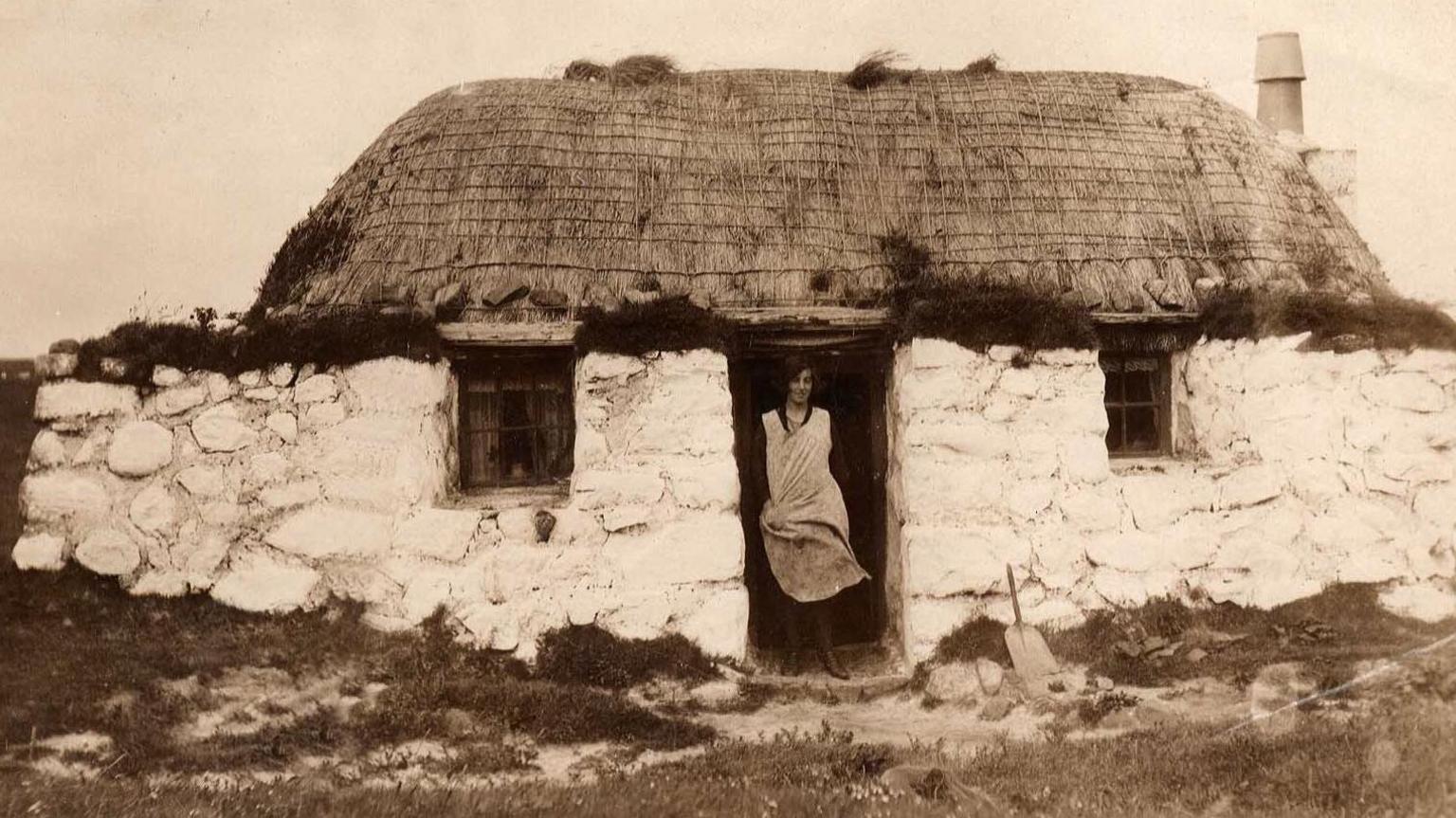 A woman standing in the doorway of thatched roof cottage