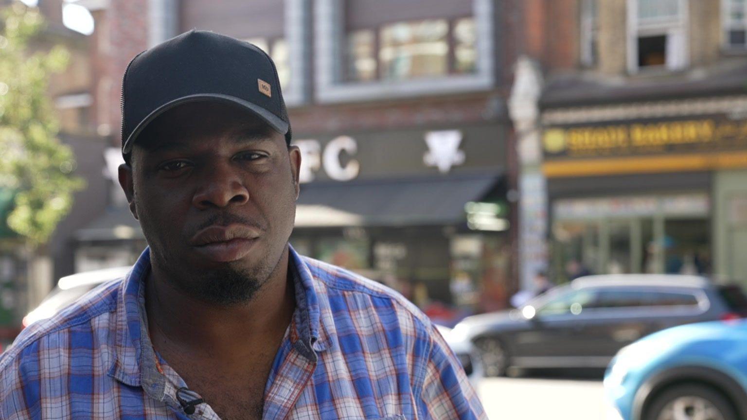 An image of Anthony King, a man wearing a baseball hat and red and blue checked shirt, standing on a street