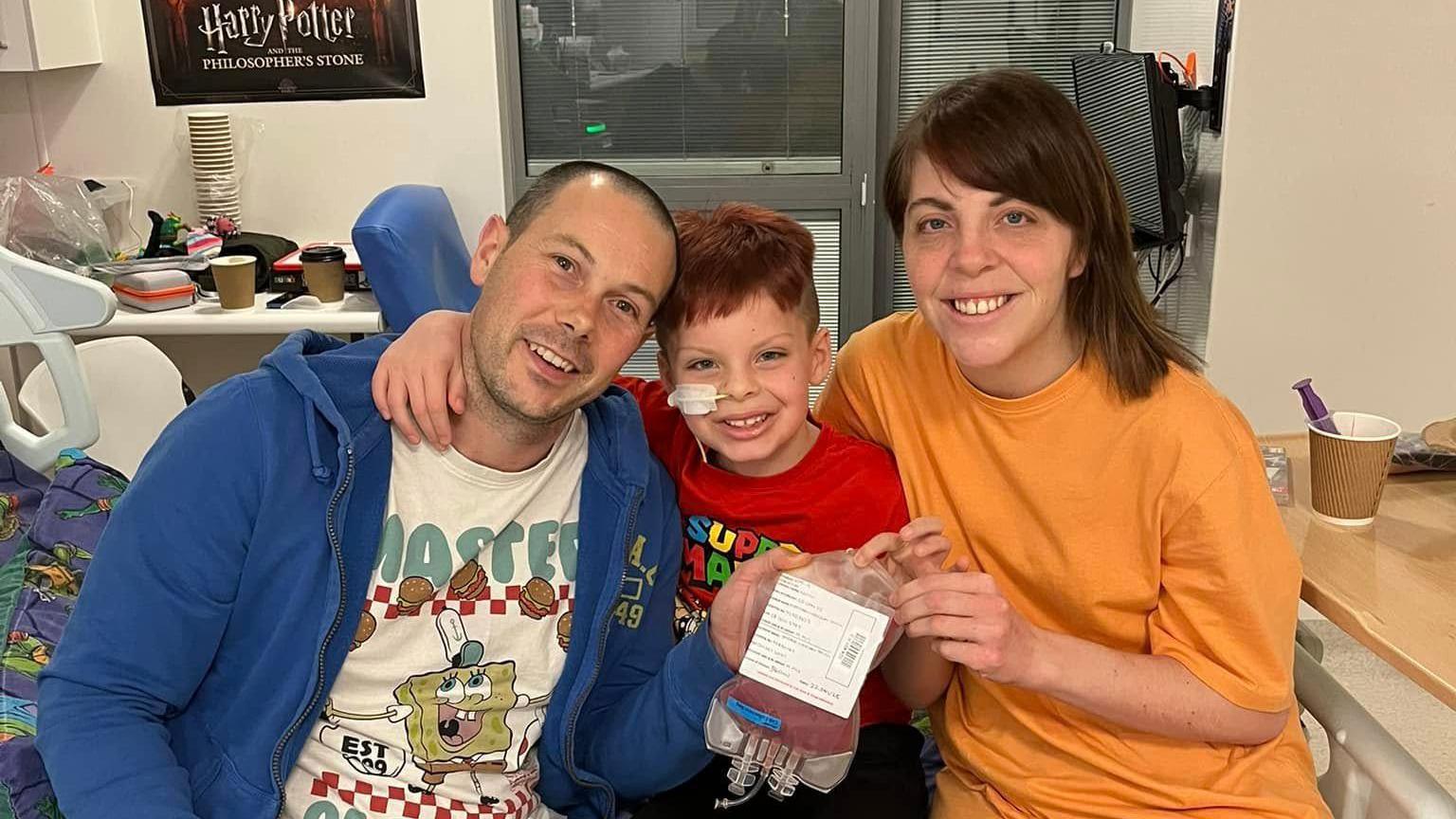 Jasper (centre) holds his new bone marrow from his dad (left) with mum Sophie (right) they all sit upon a hospital bed and smile at the camera. 