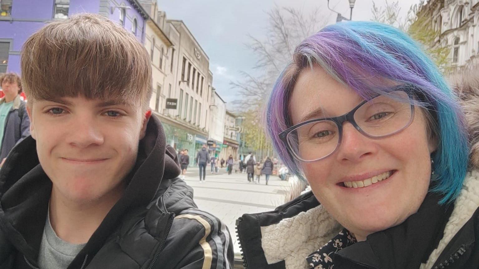 Becci Smart taking a selfie with Zak on a pedestrianised city centre street. Zak is on the left and his light brown hair is brushed forward so his fringe covers his forehead. He has a light grey t-shirt on underneath a black Adidas puffa jacket. Becci has a bob-cut hairstyle which is purple and blue. She is wearing blue-rimmed glasses and a black coat over a black dress with white flowers on. People can be seen walking down the street behind them between various shop fronts. It is a head and shoulders shot of the two of them.