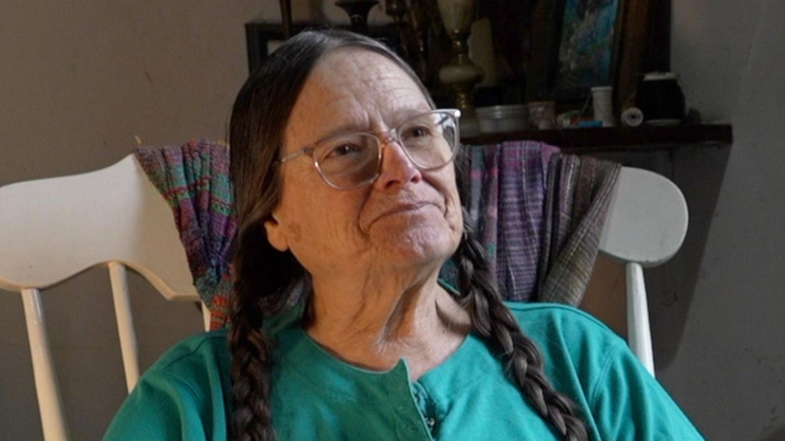 A woman with long brown hair in two plaits is sitting in a rocking chair. She is wearing a green top and glasses