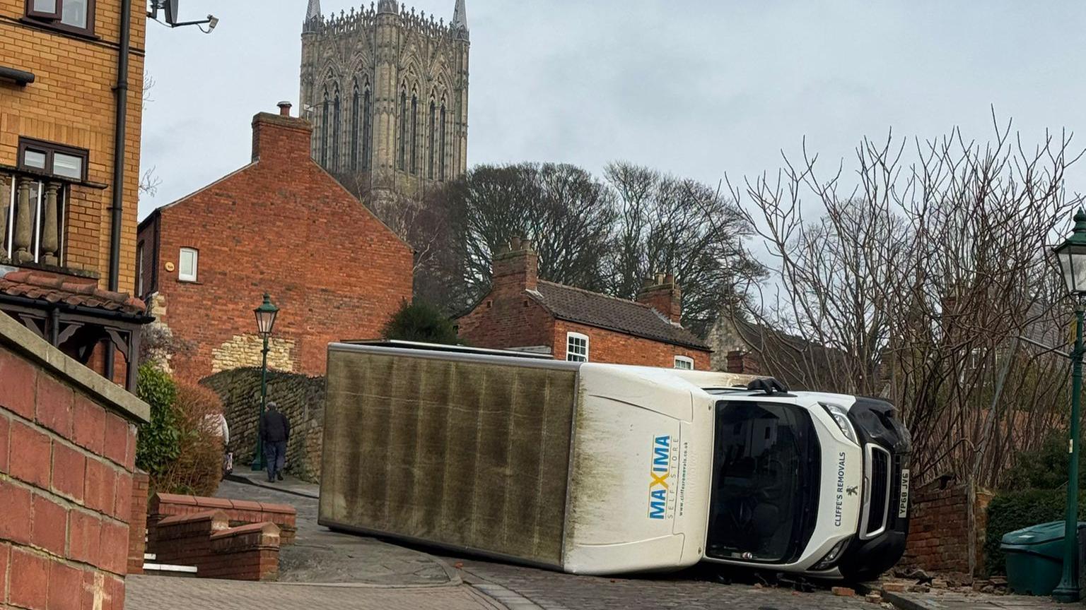 An overturned white van on a steep cobbled street. Houses can be seen surrounding the overturned van and the cathedral tower in the background. Bricks have come off a damaged wall next to where it fell.