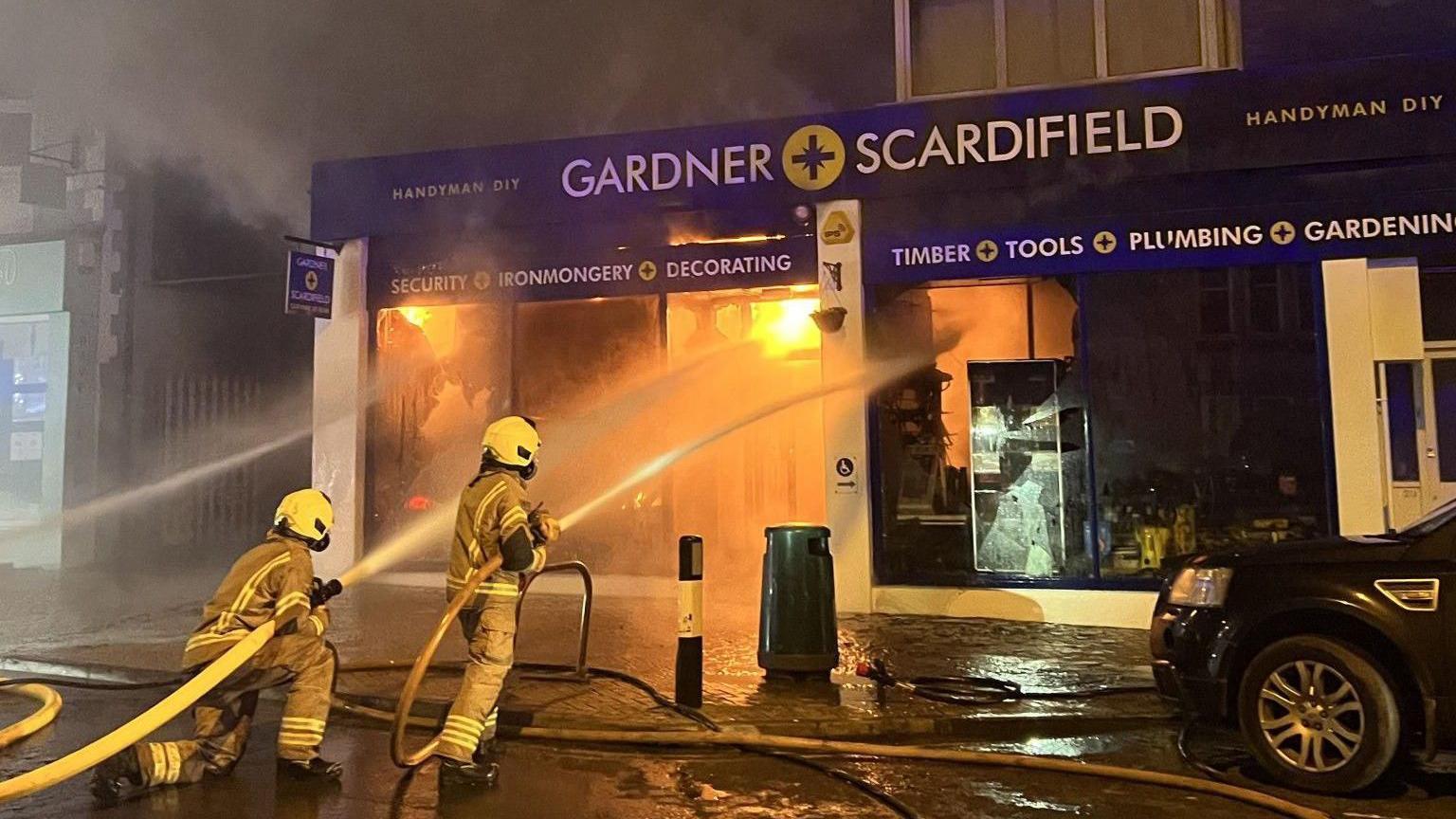 Two firefighters spraying water at a building. The building has a blue shop front and flames on the inside.