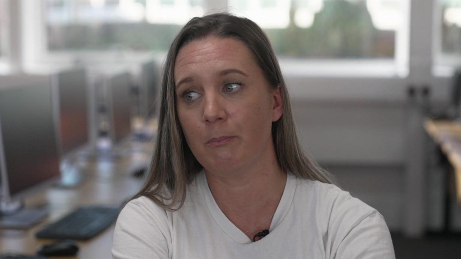 Becky Dustan sits in a classroom with a bank of computers behind her. She has a white t-shirt on and is looking to her right. She has fair straight hair which goes past her shoulders.