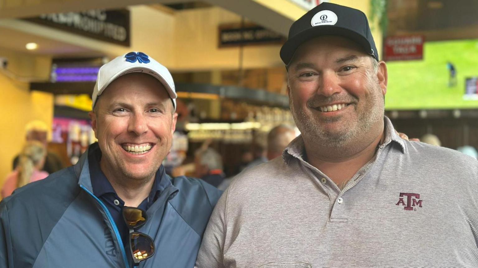 Two men are standing in a bar. The man on the left is wearing a white cap, navy polo and blue jacket, he has a pair of sunglasses hanging from his shirt. The man on the right is wearing a black cap and a red and grey polo. Behind them is a bar with a large screen. It has golf on it.