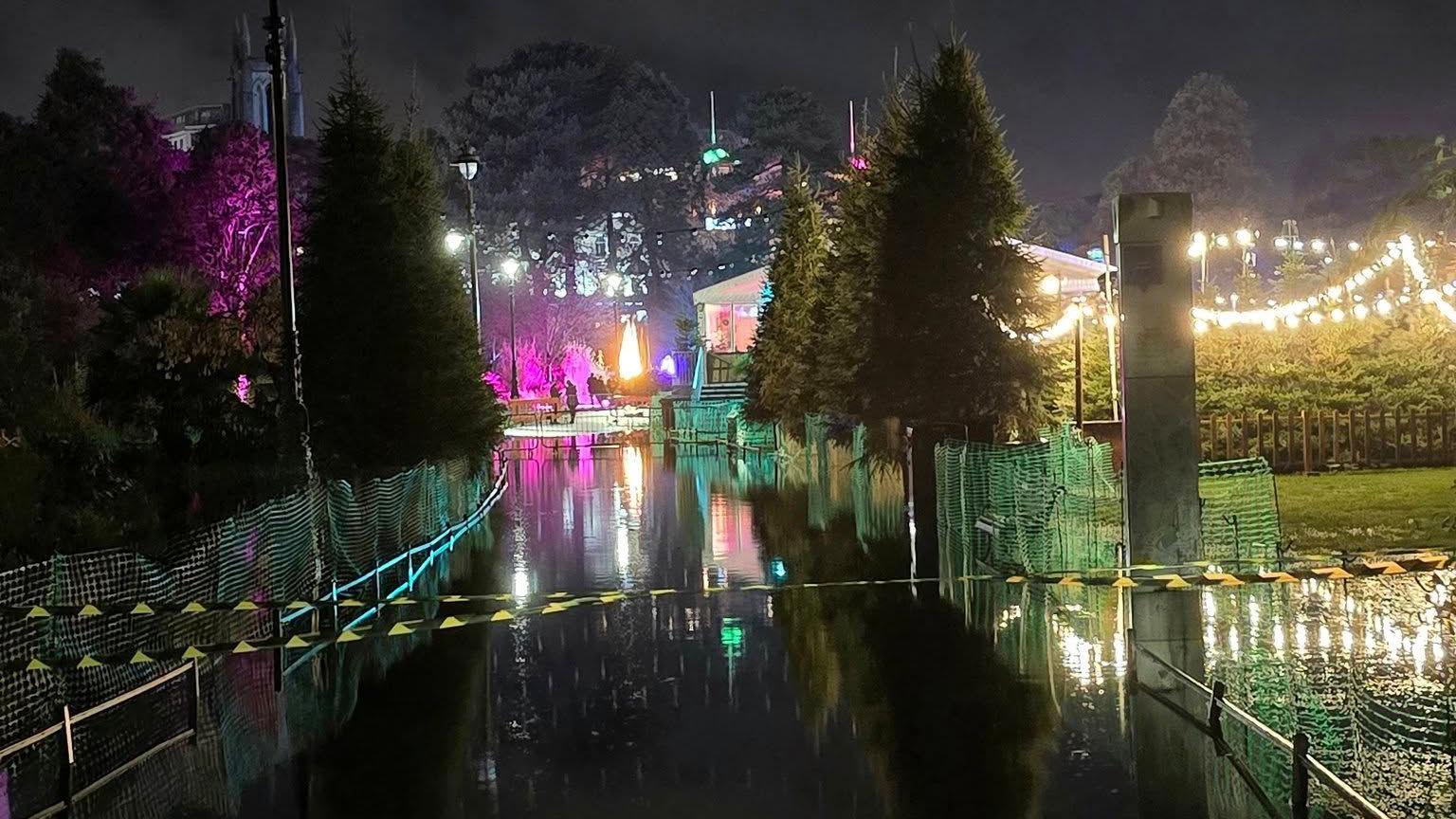 A flooded lower gardens, taped off with yellow and black tape. In the distance is the temporary ice rink which is lit and other unaffected illuminations, including trees and string lights.
