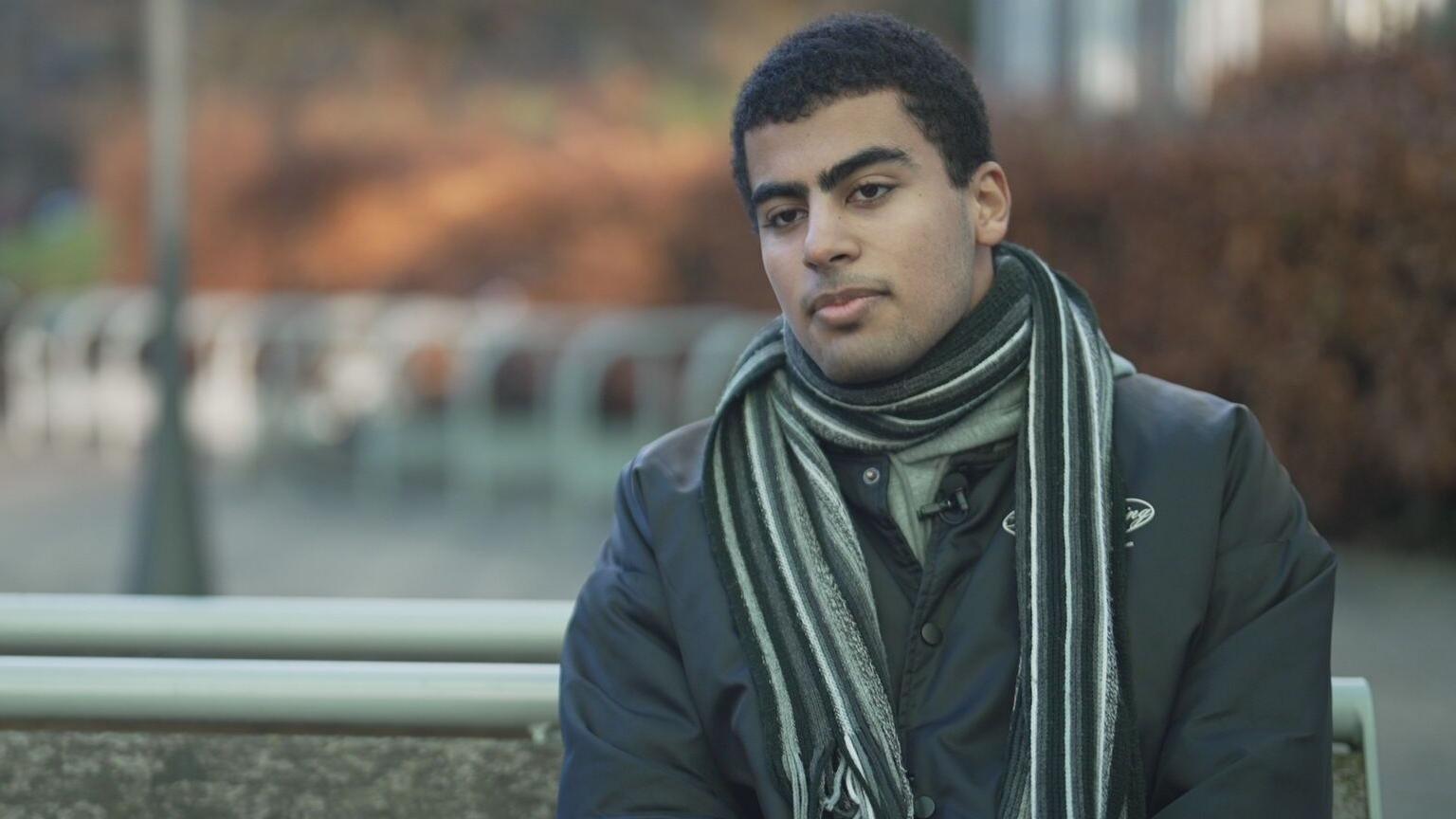 A man with short dark hair, wearing a black jacket and grey scarf, sits on a bench outdoors. He is looking forwards towards the camera. 