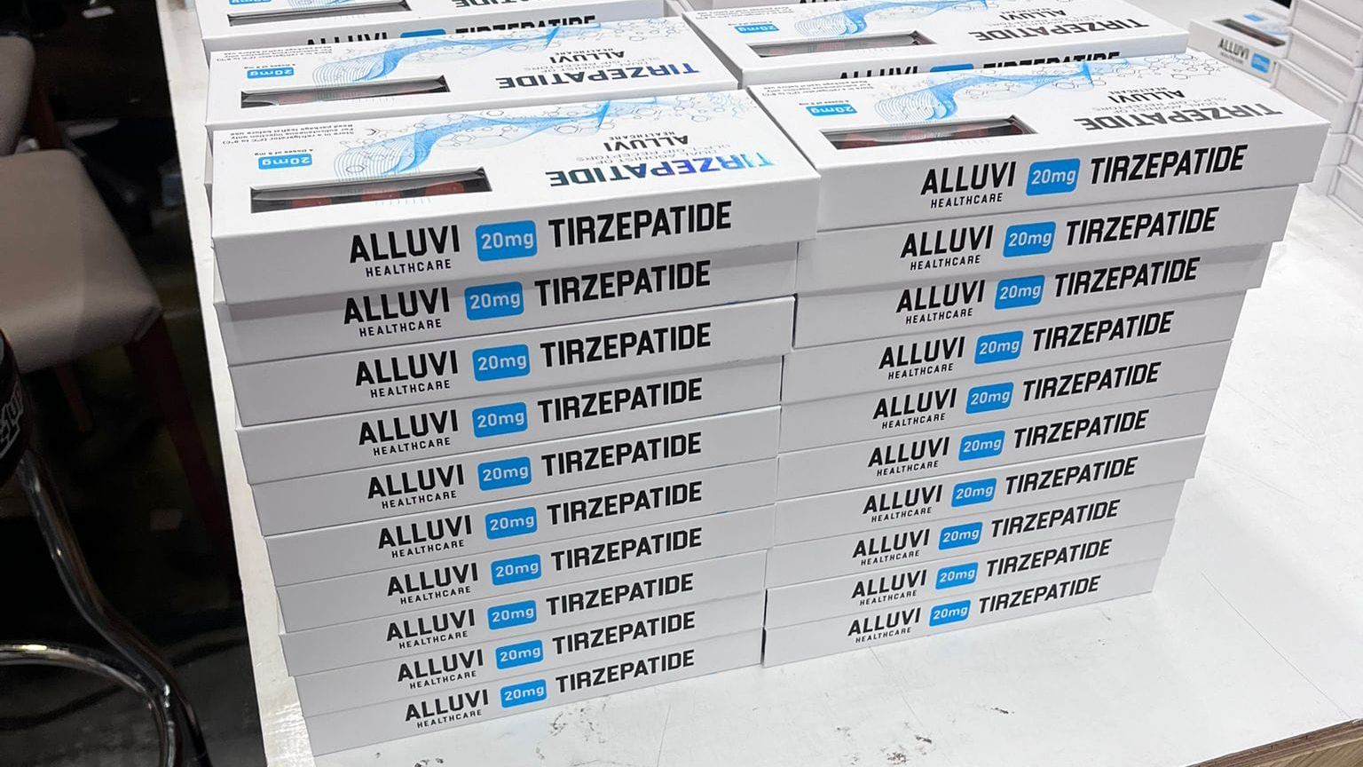 Boxes of illegal Alluvi Trizepatide drugs piled on a table. The white boxes are oblong, with blue and black writing and patterns on them. They are stacked in piles of 10, with six stacks visible in the foreground and more to the right.