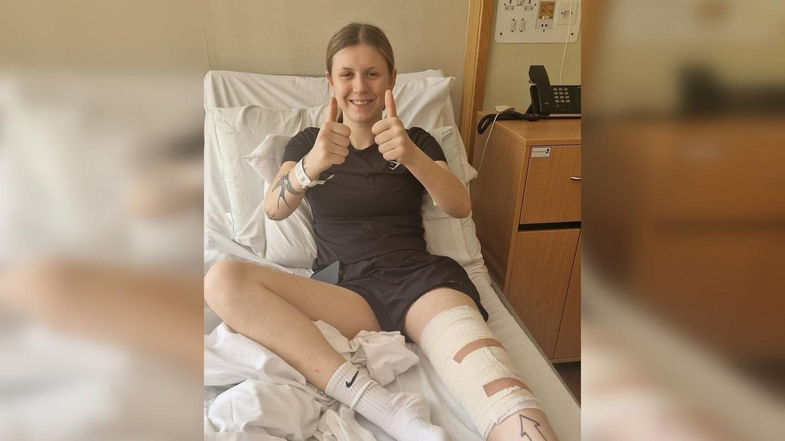 Jess yn gorwedd ar wely mewn ysbyty yn gwenu ac yn dal ei ddau bys bawd lan. Mae'n gwisgo crys T a siorts ddu a chlytiau o gwmpas ei phen-glin.