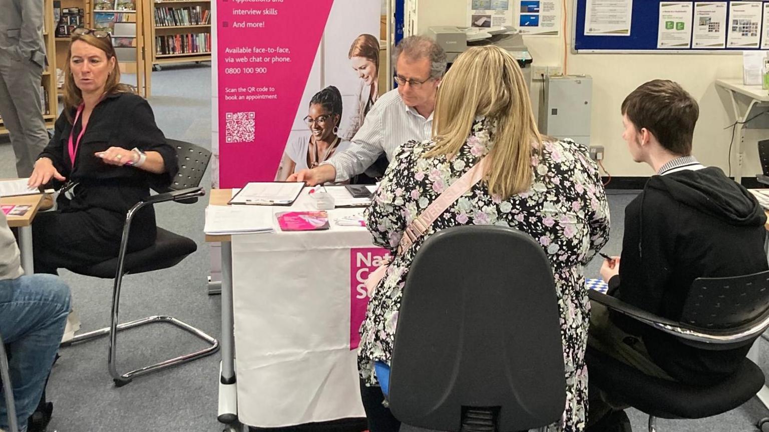 A woman and a man sit opposite another man in a white shirt, who is picking up a clipboard. They are all sat at a table. 