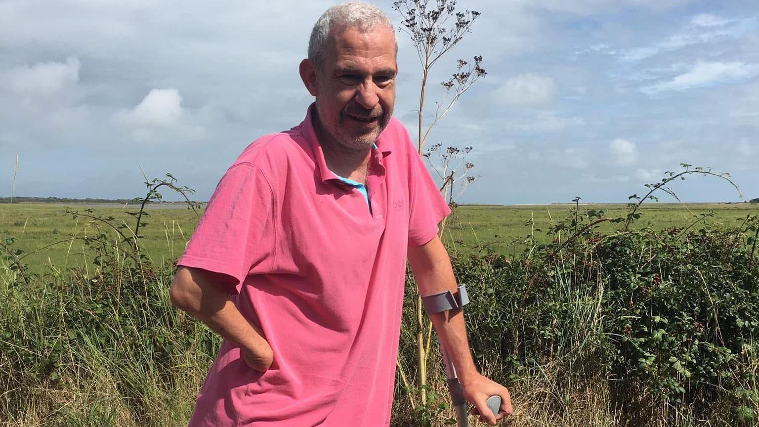 Steven Lacey standing against a countryside backdrop. Steven is standing with a crutch
