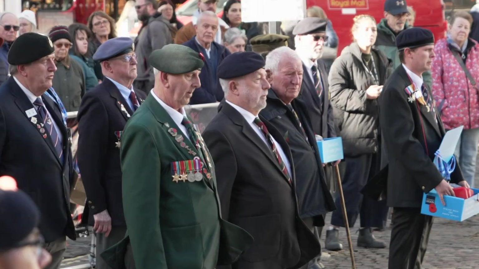 The image shows a group of people standing outdoors in Truro city centre. Several individuals in the foreground are wearing military-style blazers, berets, and medals. One person is holding a box with items inside and another carries a walking stick. Behind them, there is a crowd of onlookers dressed in casual clothing. A bright red Royal Mail van is visible in the background.