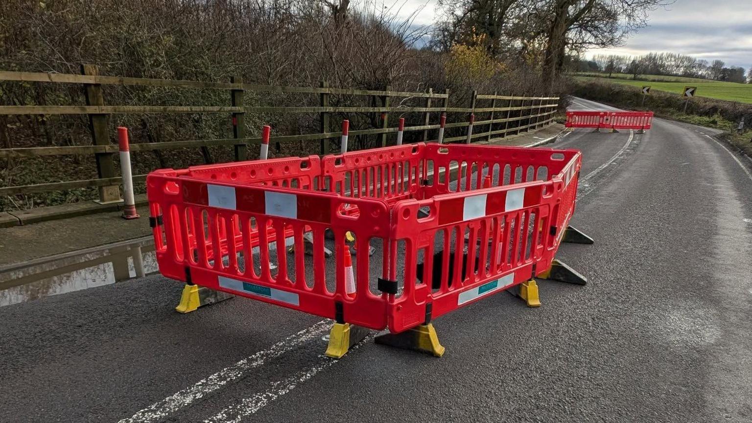 The road with barriers around the sinkhole. Some road cones can be seen as well. The surface is damp. It's a cloudy day.