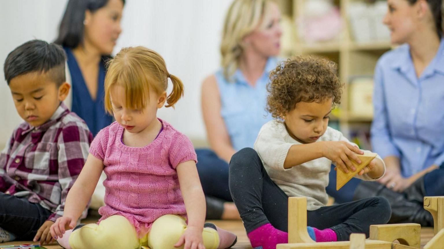 Three young children, two boys and a girl, playing with building blocks in a library with their parents sat behind them.