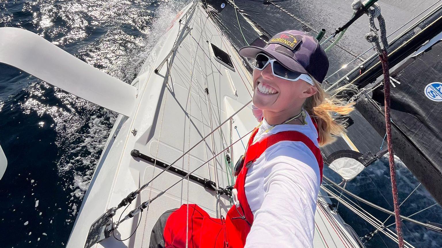 Meg Niblett is wearing a white long sleeved top, red overalls, a cap, white rimmed sunglasses and is wide smiling at the camera as she takes a selfie from the deck of a white yacht with the blue black ocean visible at the sides