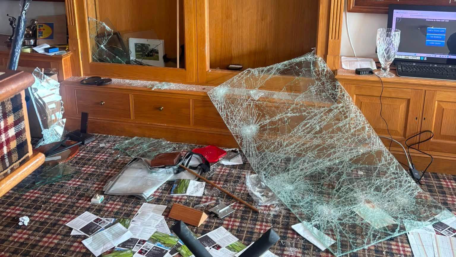 Significant damage to a wooden display cabinet following an apparent impact, with shattered glass panels, debris scattered across the floor, and contents displaced throughout the room.